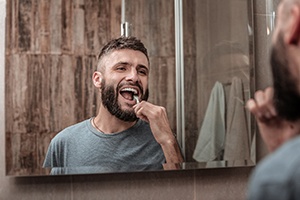 A man with a beard stands at his bathroom mirror and brushes his teeth